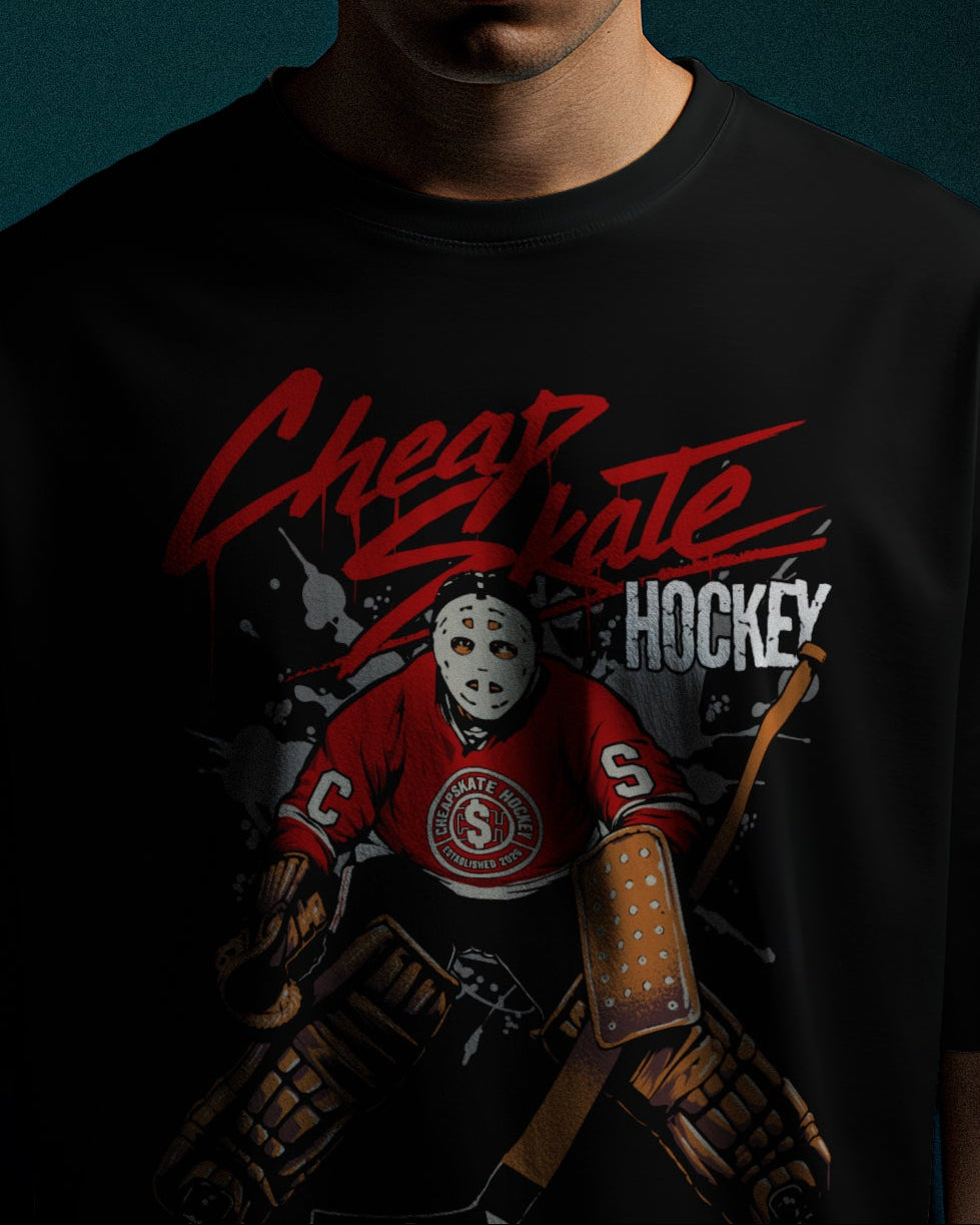 A man wearing a beige baseball cap and a black graphic t-shirt with a red and white hockey design, standing against a dark teal background.