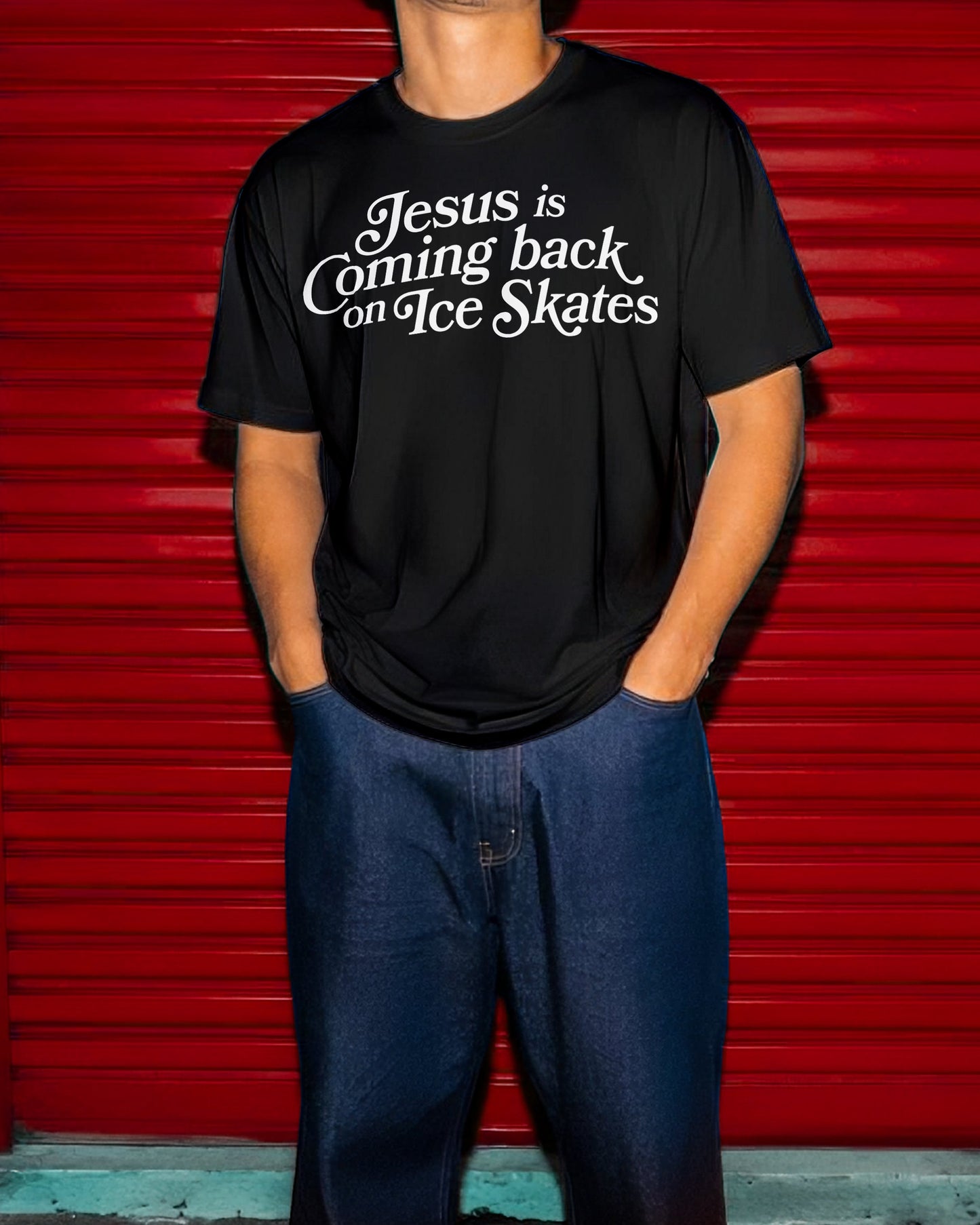 A person wearing a black graphic t-shirt with the white text 'Jesus is Coming back on Ice Skates' and blue jeans, standing in front of a bright red metal shutter.